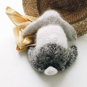 May include: A gray and white knitted bunny rabbit toy with a fluffy tail. The bunny is lying on its back with its paws in the air.