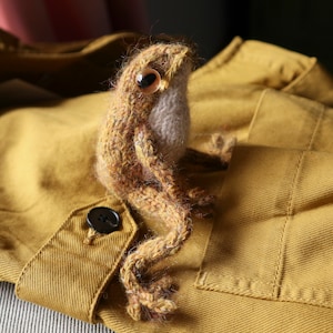 May include: A brown and beige knitted frog toy with a button eye, sitting on a mustard yellow fabric background.