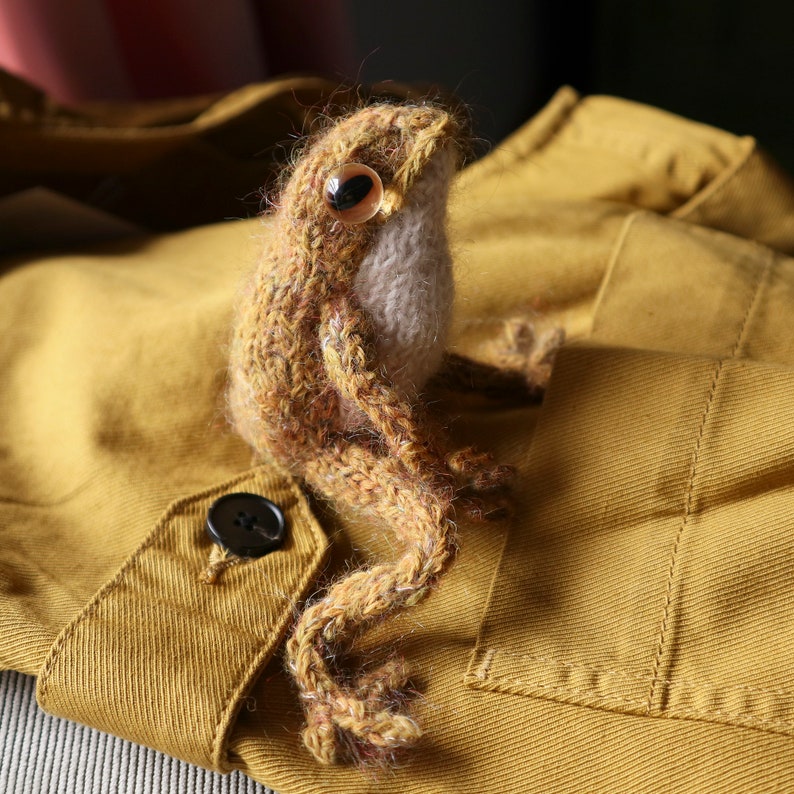 May include: A brown and beige knitted frog toy with a button eye, sitting on a mustard yellow fabric background.