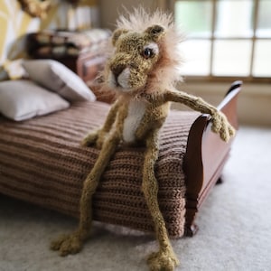 Könnte beinhalten: Eine handgestrickte Löwenpuppe mit einem beigefarbenen Körper, einer weißen Brust und einer flauschigen, hellbraunen Mähne. Der Löwe sitzt auf einem Miniaturbett mit einer gestrickten Decke. Die Puppe hat eine entspannte Haltung, mit einem Arm auf der Bettkante.
