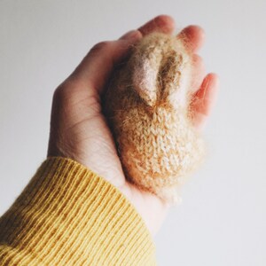 Puede incluir: Una mano sosteniendo un peque&ntilde;o juguete de conejo tejido. El conejo es beige con una nariz y orejas de color rosa claro. La mano lleva un su&eacute;ter amarillo.