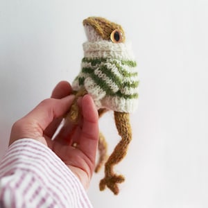 May include: A knitted frog wearing a white and green striped sweater. The frog has brown yarn legs and a brown yarn body. The frog is being held by a hand.