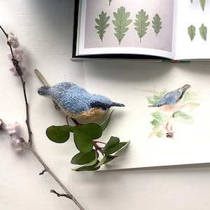 May include: A blue and brown knitted bird figurine perched on a branch of green leaves. The bird is positioned above a watercolor painting of a similar bird on a branch.