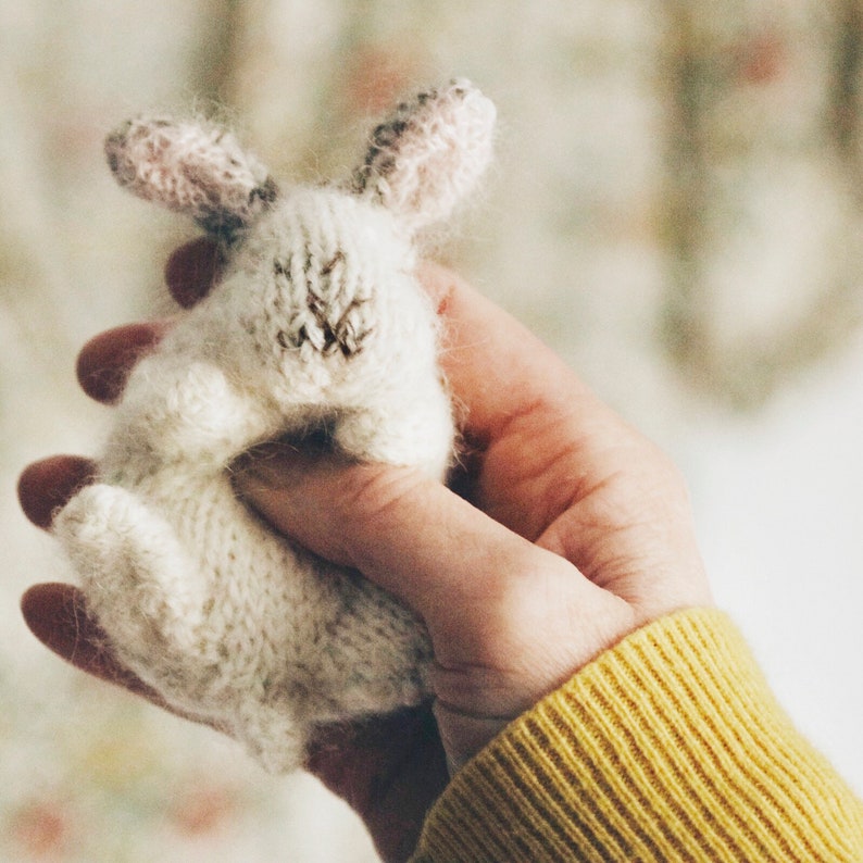 Puede incluir: Un juguete de conejo blanco de punto con orejas rosas, sostenido en una mano que lleva un su&eacute;ter amarillo.