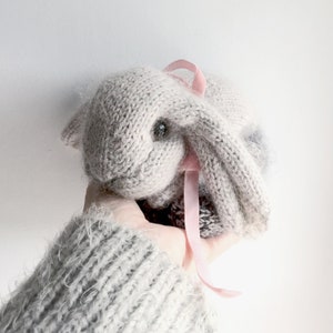 May include: A hand holding a gray knitted bunny with a pink ribbon tied around its neck.