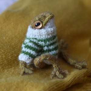 May include: A small knitted frog wearing a green and white striped sweater. The frog has a brown body and large, round eyes.