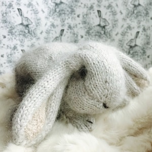 May include: A gray knitted bunny rabbit toy with long floppy ears. The bunny is lying on a white fluffy surface.