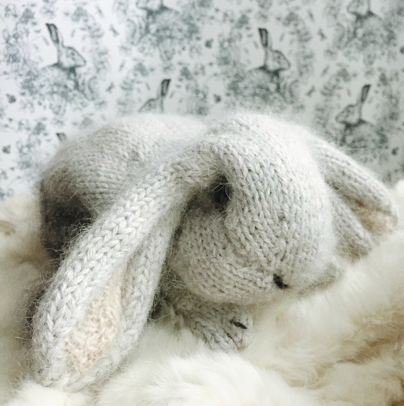 May include: A gray knitted bunny rabbit toy with long floppy ears. The bunny is lying on a white fluffy surface.