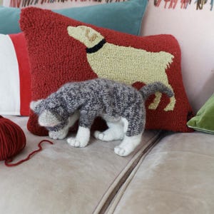 Peut inclure: Une figurine de chat tricoté gris et blanc, avec des pattes blanches et un nez rose, devant un coussin rouge avec un motif de chien. Une pelote de laine rouge est à gauche. Autres coussins en arrière-plan.