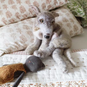 Könnte beinhalten: Handgestrickte Wolfspuppe in Hellgrau und Creme, sitzend auf einem Bett mit gemusterten Kissen. Der Wolf hat eine schwarze Nase und braune Augen. Eine kleine Puppe mit brauner Weste liegt daneben.