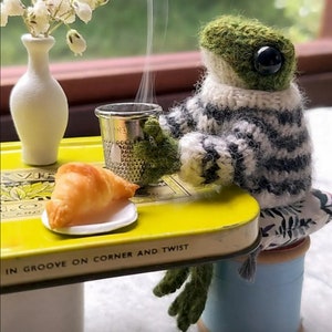 May include: A green knitted frog wearing a black and white sweater sits at a yellow table with a croissant and a metal thimble. The table has the text "in groove on corner and twist" printed on the side.