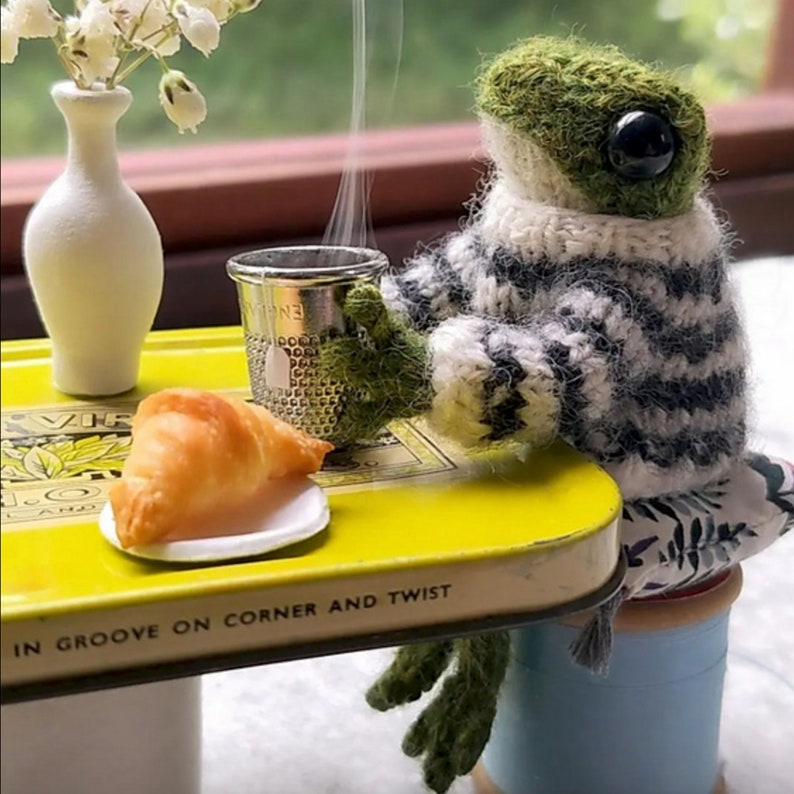May include: A green knitted frog wearing a black and white sweater sits at a yellow table with a croissant and a metal thimble. The table has the text "in groove on corner and twist" printed on the side.