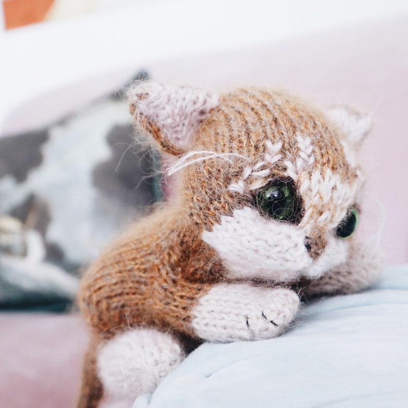 May include: A brown and white knitted cat toy with green eyes. The cat is sitting on a light blue surface.