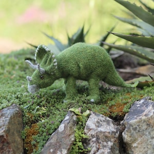 Può includere: Un giocattolo di dinosauro triceratopo di colore verde lavorato a maglia con corna grigie e una superficie testurizzata. Il giocattolo è seduto su un letto di muschio e rocce.