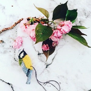 Op de afbeelding: Een gebreide blauwe vogel met gele accenten zit op een wit marmeren oppervlak. Roze kersenbloesems met groene bladeren zijn rond de vogel verspreid.