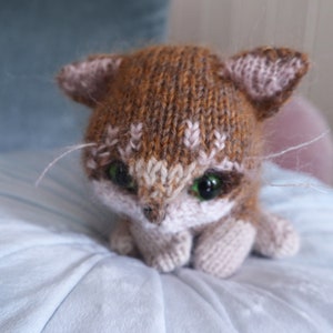 May include: A brown and white knitted cat toy with green eyes. The cat is sitting on a light blue surface.
