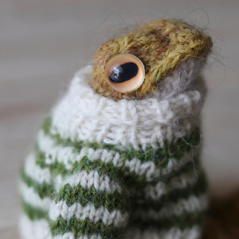 May include: A knitted frog wearing a green and white striped sweater with a white turtleneck. The frog has a large brown eye with a black pupil.