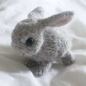 May include: A gray knitted bunny rabbit toy with black button eyes. The bunny is sitting on a white surface.