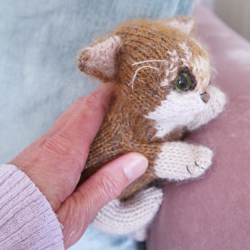 May include: A brown and white knitted cat toy with green eyes. The cat is being held in a hand.