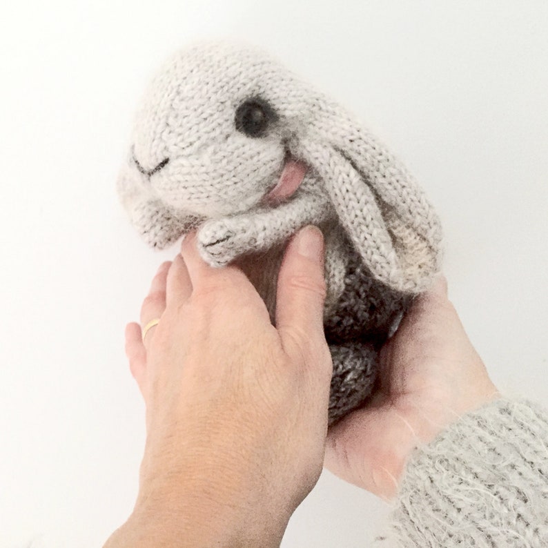May include: A hand-knitted gray bunny rabbit toy with black eyes. The bunny is being held by two hands.