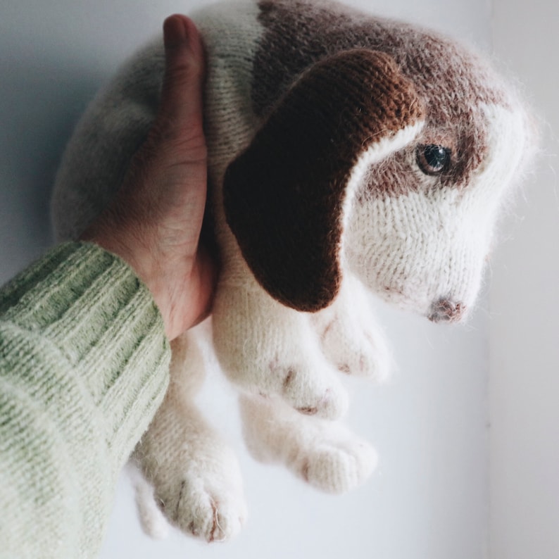 May include: A hand holding a knitted brown and white dog toy. The toy has a long snout and floppy ears.