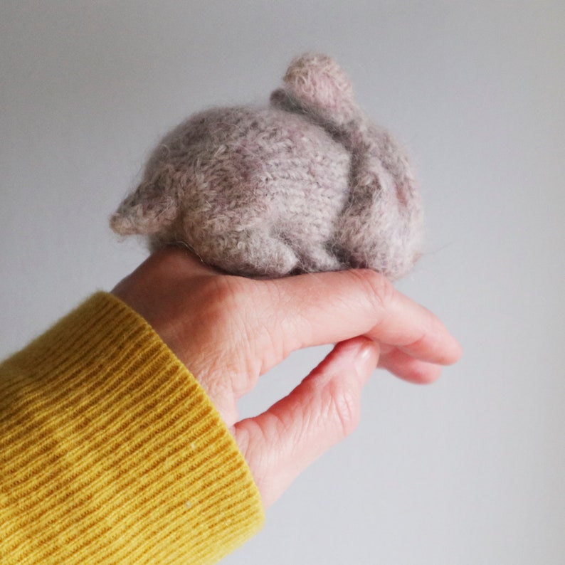 Puede incluir: Un peque&ntilde;o juguete de conejo tejido a mano en color morado claro. El conejo est&aacute; acurrucado en posici&oacute;n de dormir.