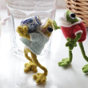 Puede incluir: Dos figuras de rana tejidas. Una rana lleva un suéter azul claro y lleva una pequeña taza de té con estampado azul y blanco. La otra rana lleva un suéter rojo con ribete blanco. Ambos tienen grandes ojos negros y extremidades verdes.