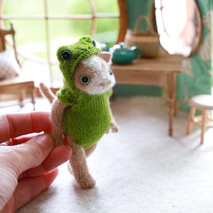 Może przedstawiać: Ręcznie wykonana miniaturowa figurka kota w zielonym kostiumie żaby. Kot ma jasnobrązowe i białe futro, z dużymi, okrągłymi oczami. Kostium żaby ma kaptur z guzikowymi oczami i zielone ciało. Figurka jest trzymana w dłoni.