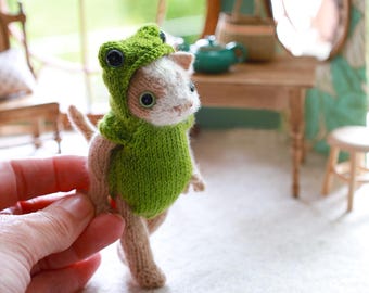 May include: A handmade, miniature cat figurine wearing a green frog costume. The cat has tan and white fur, with large, round eyes. The frog costume includes a hood with button eyes and a green body. The figurine is held in a person's hand.