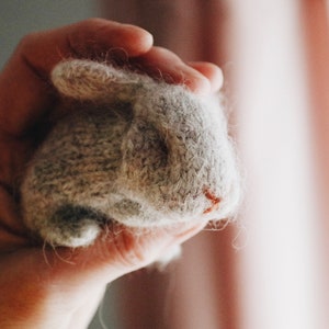 Puede incluir: Una mano sosteniendo un peque&ntilde;o conejo de punto gris. El conejo tiene una textura esponjosa y una nariz rosa.