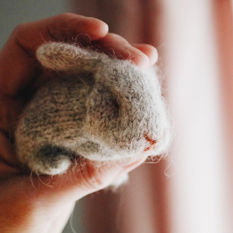 Puede incluir: Una mano sosteniendo un peque&ntilde;o conejo de punto gris. El conejo tiene una textura esponjosa y una nariz rosa.