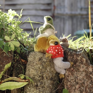 Peut inclure: Deux figurines de grenouilles en tricot, l'une verte et l'autre brune, assises sur une citrouille en velours jaune. La grenouille brune porte un pull rouge &agrave; pois blancs et un pantalon gris.