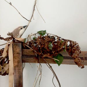 May include: A small brown and white knitted bird perched on a branch with dried brown leaves and a tan ribbon.