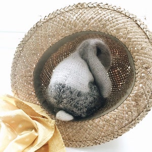 May include: A knitted gray and white bunny rabbit toy is tucked inside a straw hat. The hat is light brown and has a woven texture.