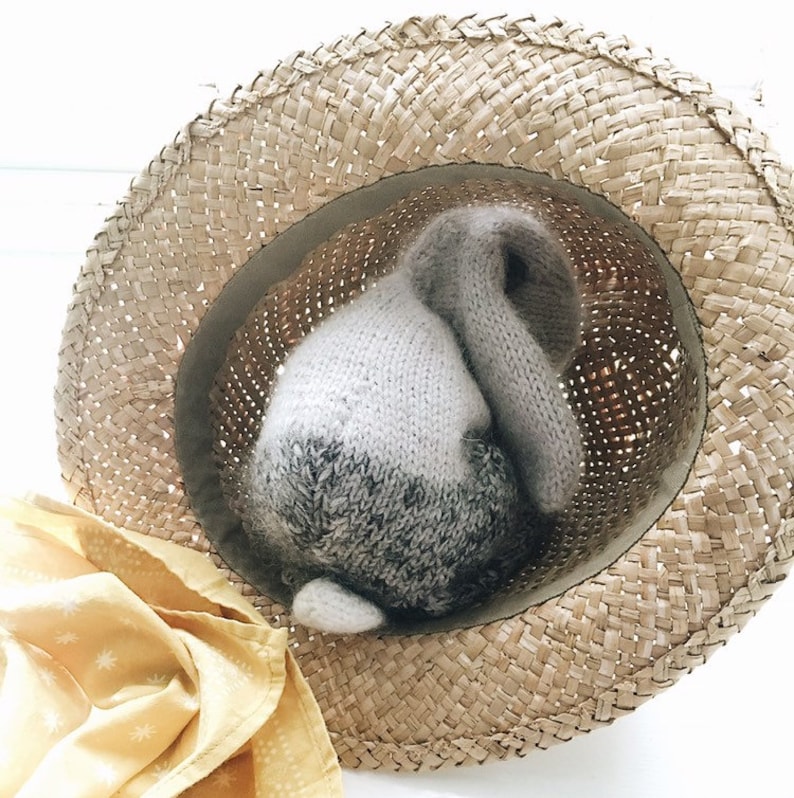 May include: A knitted gray and white bunny rabbit toy is tucked inside a straw hat. The hat is light brown and has a woven texture.