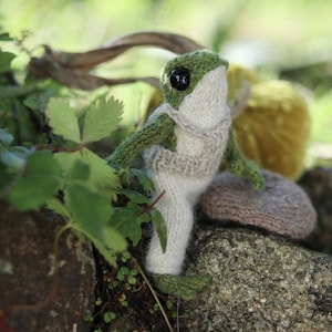 Peut inclure: Un jouet en forme de grenouille tricot&eacute; portant un pull et un pantalon blancs. La grenouille est assise sur un rocher avec de la v&eacute;g&eacute;tation verte en arri&egrave;re-plan.