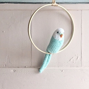 May include: A light blue knitted bird with white accents hangs from a wooden embroidery hoop. The bird is a popular craft project and home decor item.