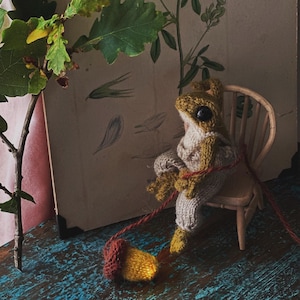 Peut inclure: Une poup&eacute;e de grenouille en tricot portant un pull brun et jaune et un pantalon rouge est assise sur une chaise en bois. La grenouille tient une ficelle rouge attach&eacute;e &agrave; un gland jaune et brun.