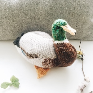 Puede incluir: Un juguete de pato silvestre tejido con un esquema de color marrón, blanco y verde. El pato está acostado sobre una superficie blanca con una rama y una pequeña planta verde en el fondo.