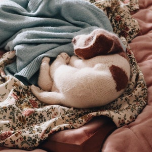 May include: A white knitted dog toy with brown patches, lying on a floral patterned fabric. The toy is curled up and has a soft, fluffy texture.