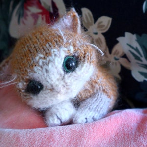 May include: A close-up of a knitted cat toy with white fur and orange accents. The cat has green eyes and is lying on a pink surface.