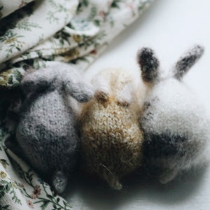 Puede incluir: Tres conejos de punto de diferentes colores: gris, beige y blanco. Los conejos est&aacute;n sentados juntos sobre una superficie blanca.