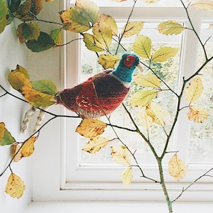 Puede incluir: Una figurita de faisán de punto con plumas rojas, azules y blancas, posada en una rama con hojas amarillas sobre un fondo blanco.