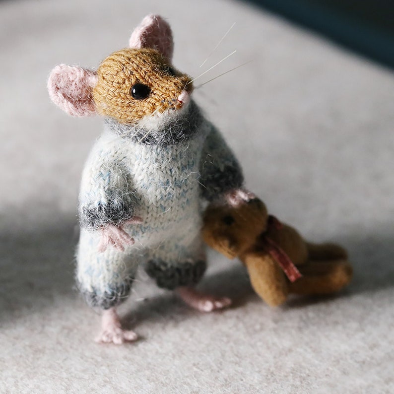 Tiny, knitted, realistic looking Mouse wearing a knitted romper suit and holding a teddy bear.