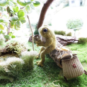 Puede incluir: Una figura de rana difusa hecha a mano en tonos verdes y beige, sosteniendo una pequeña taza. Está sentada en un tronco con una cesta de mimbre cerca, en una escena exterior en miniatura con musgo verde y follaje.