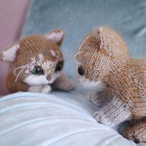 May include: Two brown and white knitted cat toys with green eyes. The cats are facing each other on a light blue surface.
