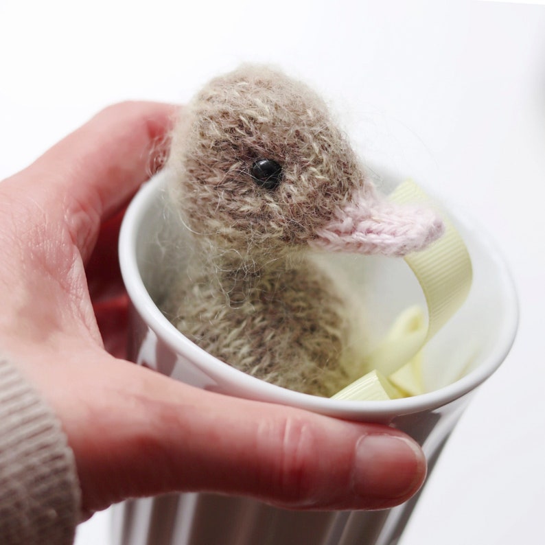 Puede incluir: Un peque&ntilde;o patito de punto con un pico rosa y pelaje marr&oacute;n est&aacute; sentado en una taza de cer&aacute;mica blanca con una cinta amarilla.
