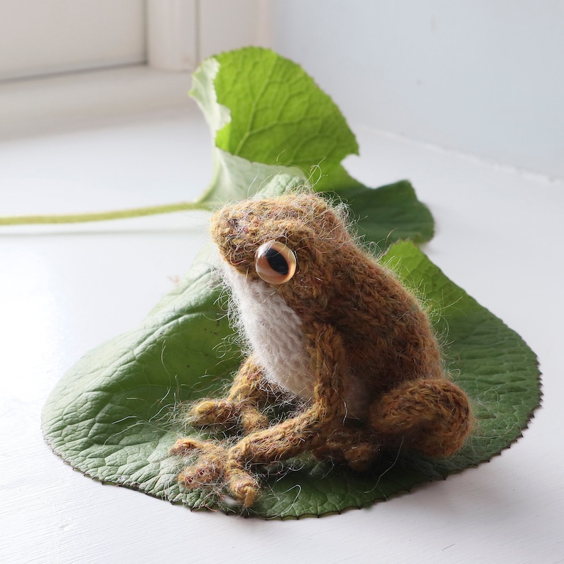 May include: A brown and white knitted frog sitting on a green leaf. The frog has large, round eyes with brown pupils.