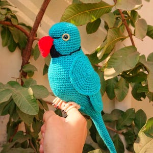 May include: A handmade turquoise crochet parrot with a red beak and black accents. The bird has a detailed, textured design and is perched on a hand. The parrot's eye is a small, black circle with a gold border. The background features green leaves.