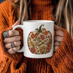 May include: A white ceramic mug featuring a central graphic of a pumpkin decorated with intricate floral patterns, autumn leaves, small coffee cups, and stacks of books in shades of orange, red, green, and brown against a white background.